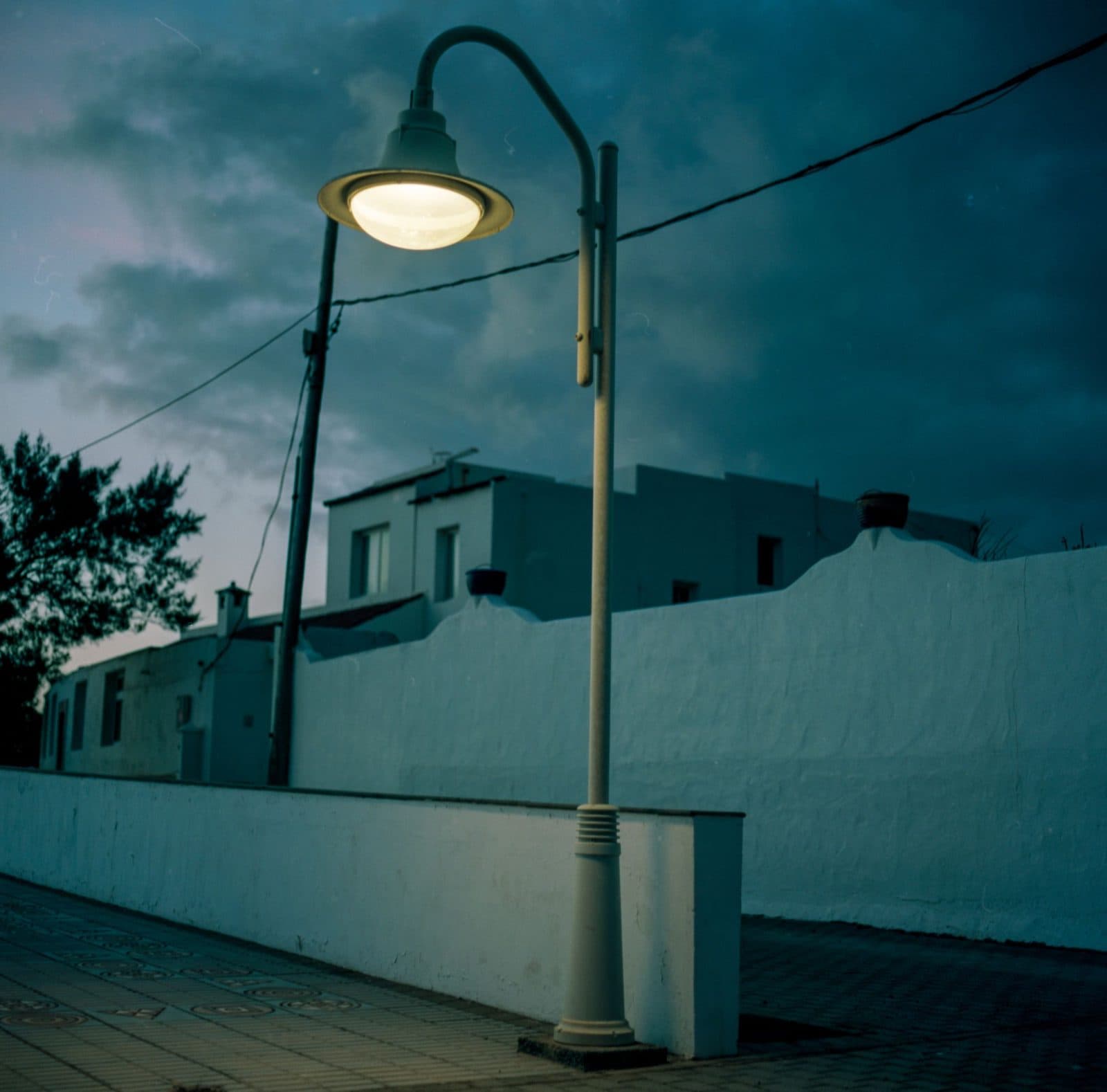 A white post-top street light on at dusk beside a white wall